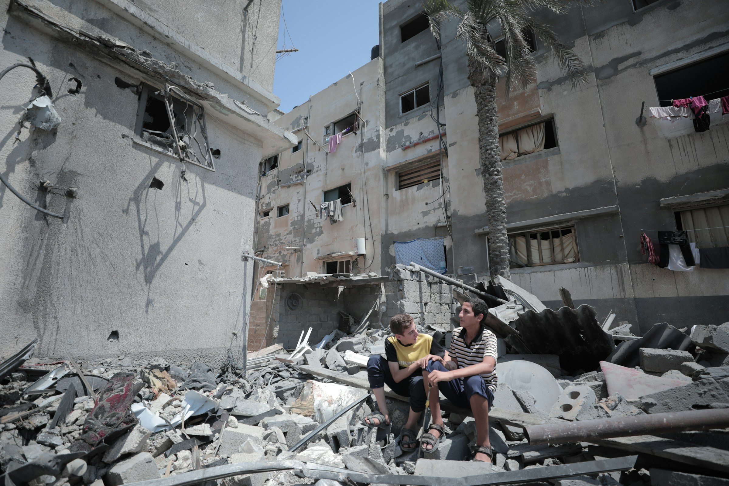 Image shows two boys sitting amidst rubble in what appears to be a war-torn or conflict-affected area. They are surrounded by debris from destroyed buildings, with the walls of a damaged structure visible behind them. The building's windows are shattered or boarded up, and laundry is hanging from some windows, suggesting that people may still be living there despite the devastation. The boys are wearing casual clothes and appear to be in conversation, possibly sharing a moment of reflection amidst the destruction. One of the boys is wearing a yellow T-shirt, and the other is wearing a striped shirt. The overall scene conveys a sense of hardship, resilience, and the impact of conflict on civilian life. The palm tree in the background adds a slight contrast to the otherwise bleak environment.