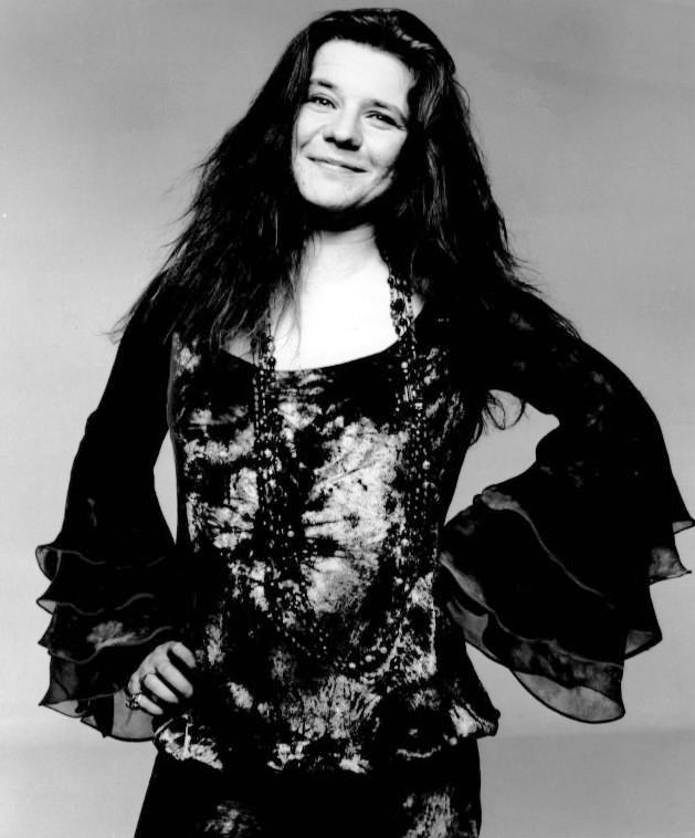 Janis Joplin in 1970 with long, wavy hair reaching past her shoulders. She is smiling gently, looking directly at the camera. She is wearing a patterned blouse with flared, ruffled sleeves. Her hands rest on her hips, and she wears long necklaces that drape over her top. The background is plain and unobtrusive, putting focus on the subject.