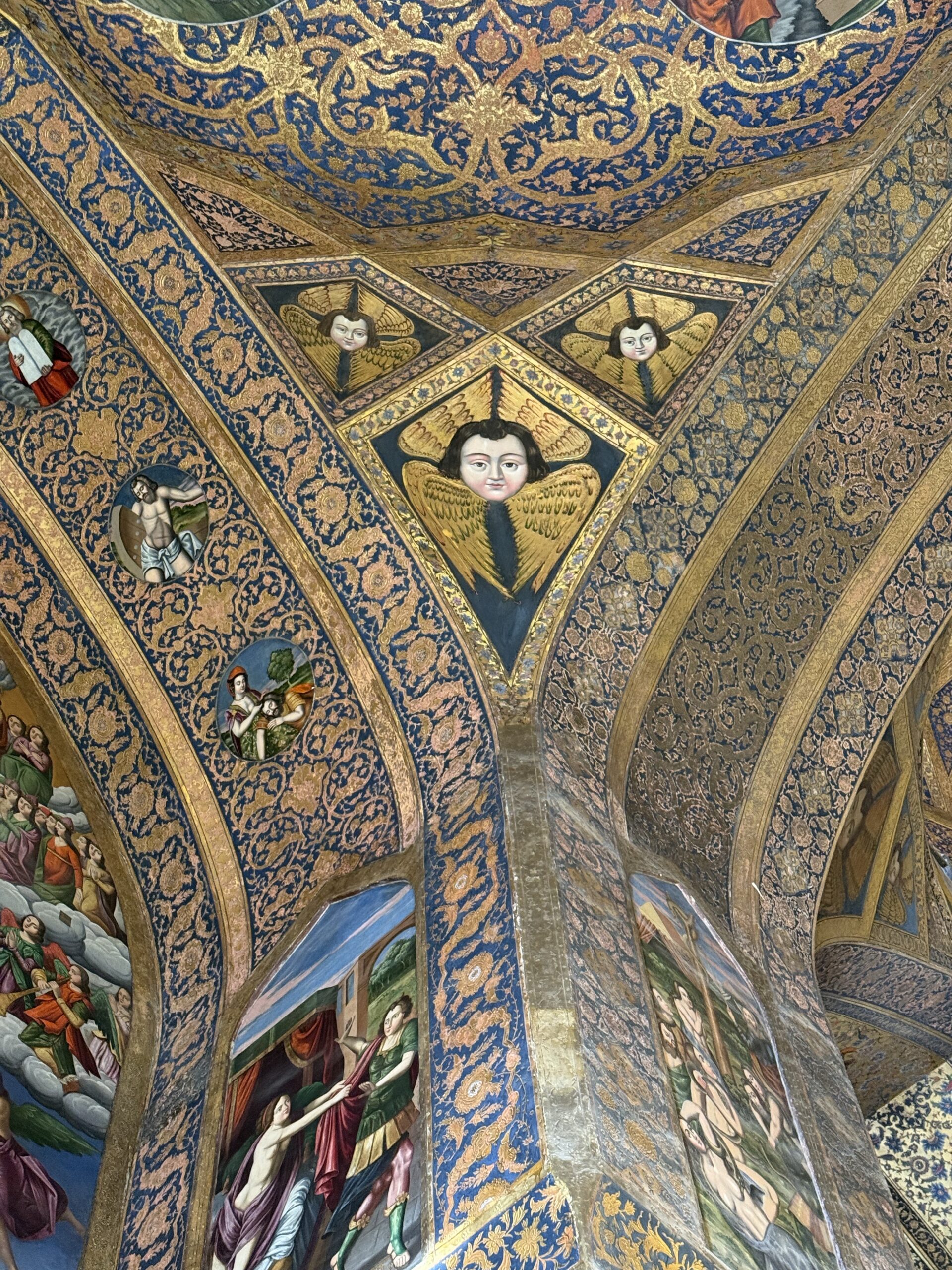 Intricately painted dome ceiling of Vank Cathedral in Isfahan, decorated with biblical scenes, golden floral patterns, and angelic faces around a central chandelier.