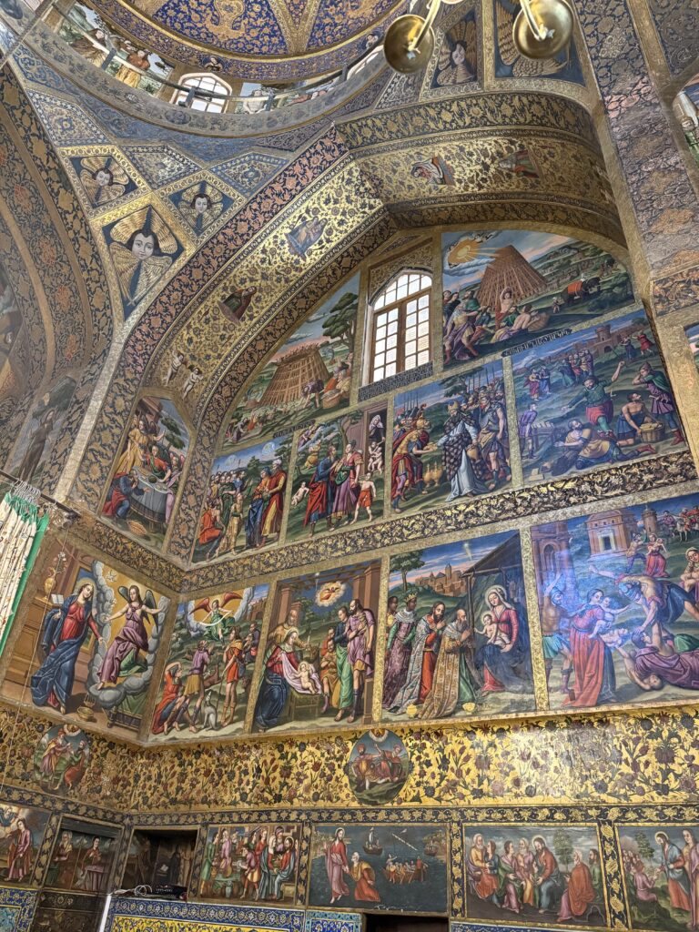 A wide view of a richly decorated wall and vaulted ceiling inside Vank Cathedral, Isfahan. The wall is covered floor-to-ceiling with colorful Christian narrative frescoes, framed by intricate blue and gold floral and geometric Safavid-style painting.