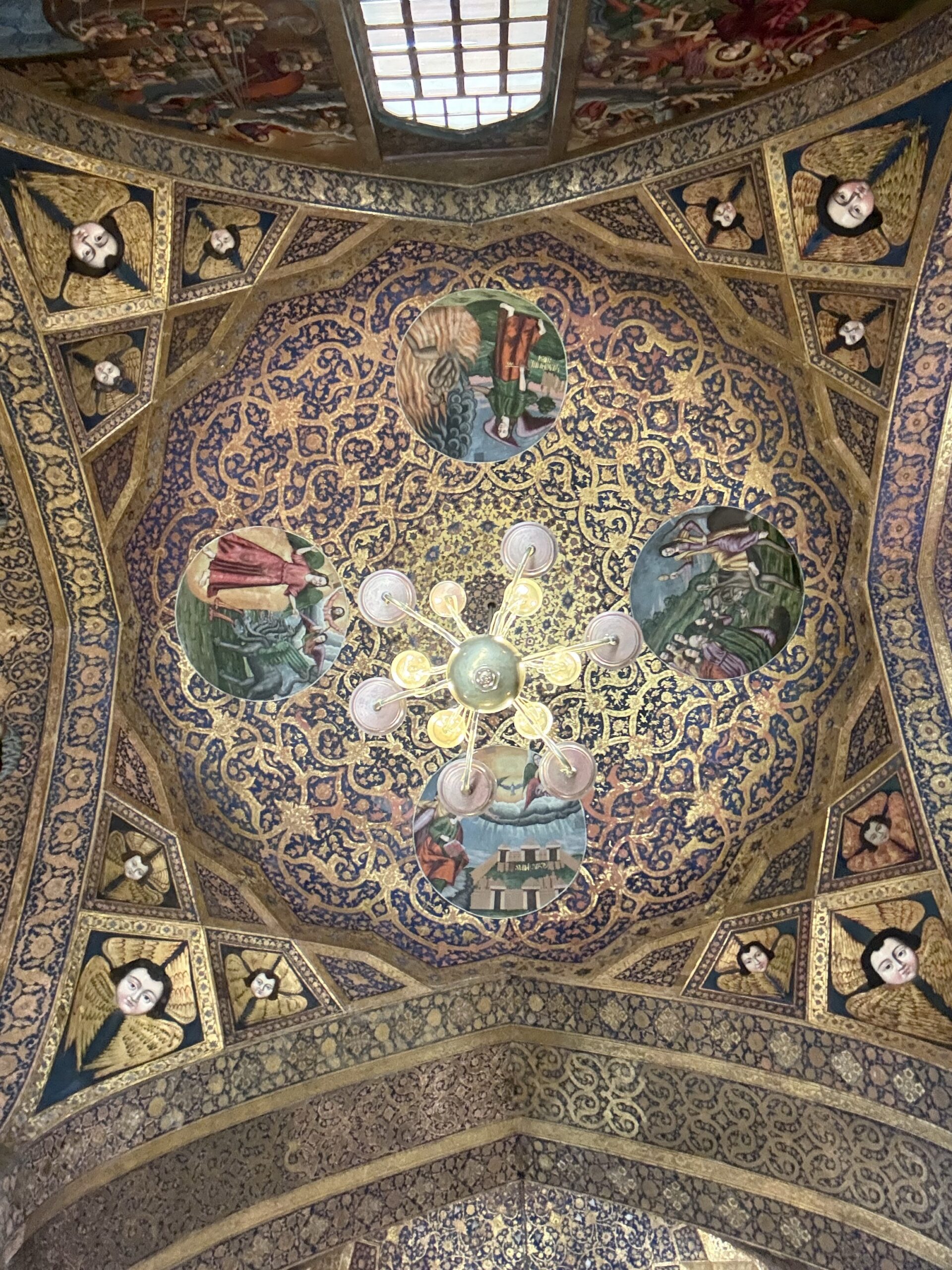 Intricately painted dome ceiling of Vank Cathedral in Isfahan, decorated with biblical scenes, golden floral patterns, and angelic faces around a central chandelier.