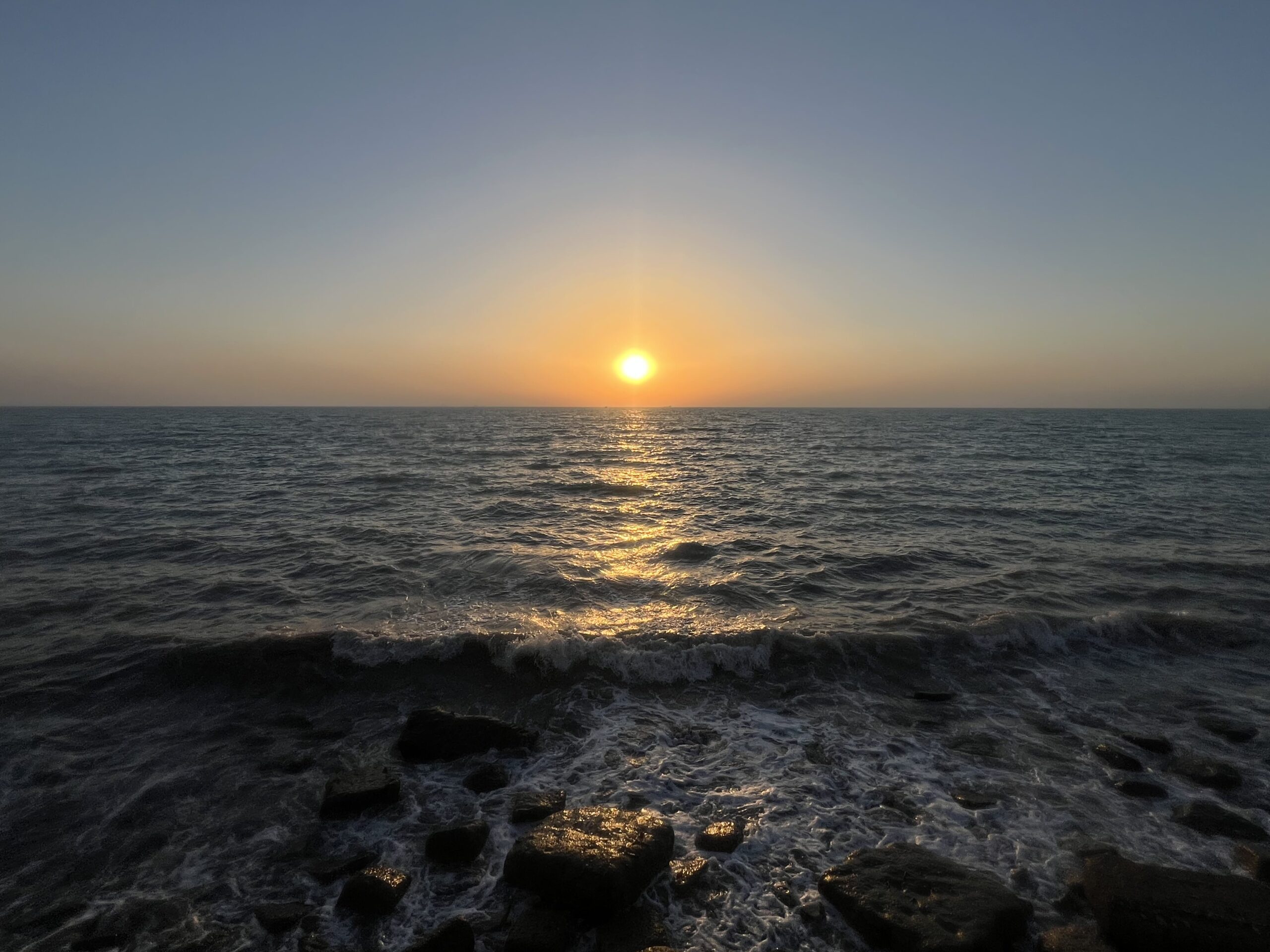 Sunset over the Persian Gulf in Bushehr, Iran, with the glowing sun just above the horizon. Warm golden light stretches across the rippling sea, reflecting toward the shore, while dark waves and rocks fill the foreground beneath a calm, hazy evening sky.